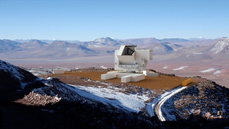 The CCAT observatory telescope facility situated on a mountain plateau with snow patches, surrounded by distant peaks under a clear blue sky.