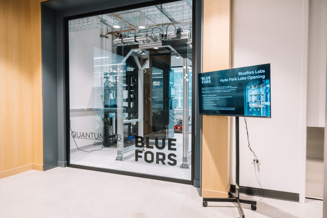 Cryogenic system visible through glass wall at the Bluefors Lab, with an opening event display screen nearby.